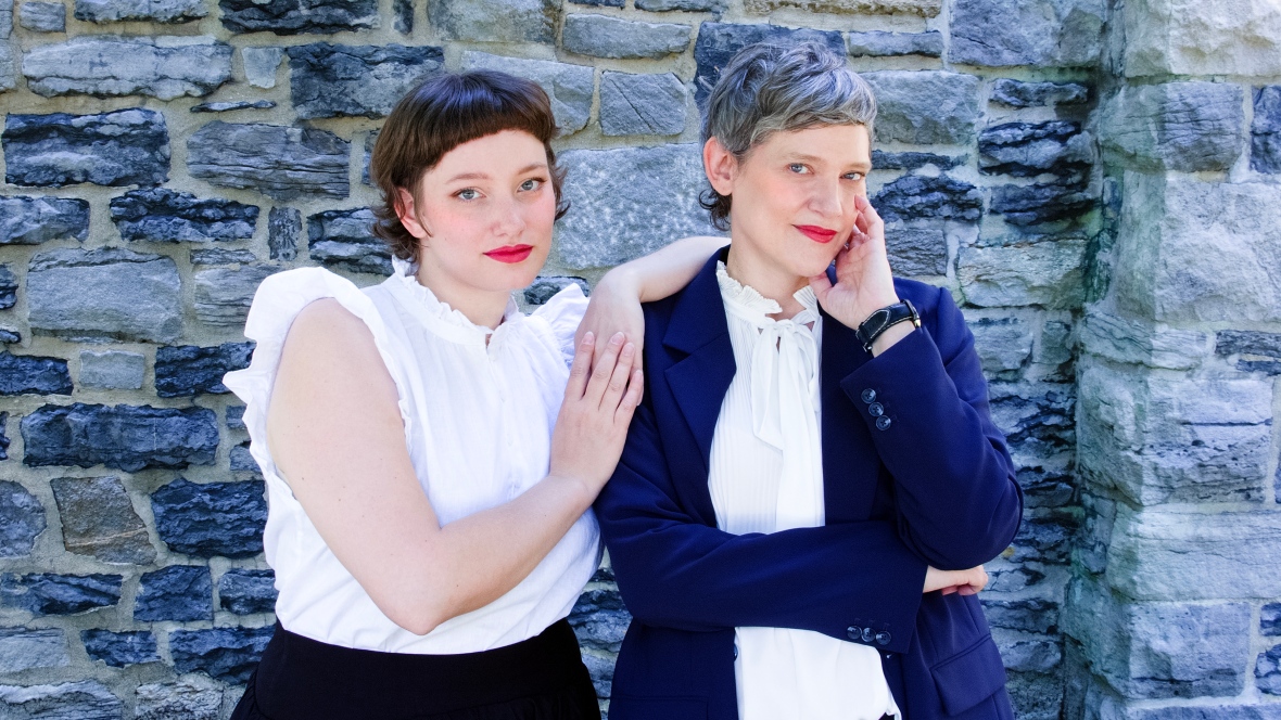 Two white women pose in front of a stone brick wall.