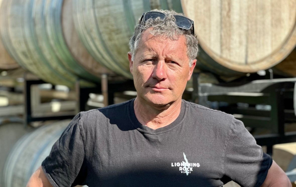A man in a black t-shirt stands in front of wood wine barrels.