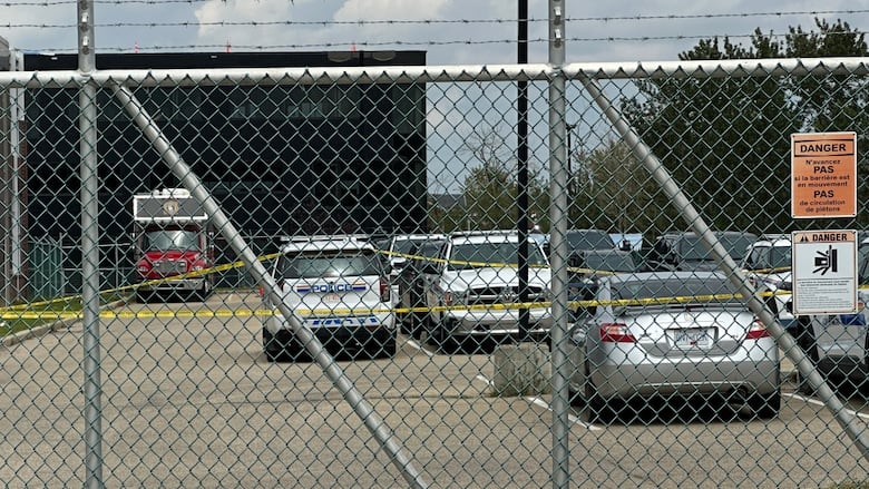 A picture of a parking lot seen through a chain link fence. Yellow police tape surrounds a set of cars. A police cruiser is one of those cars. A large two-story building is seen in the background.