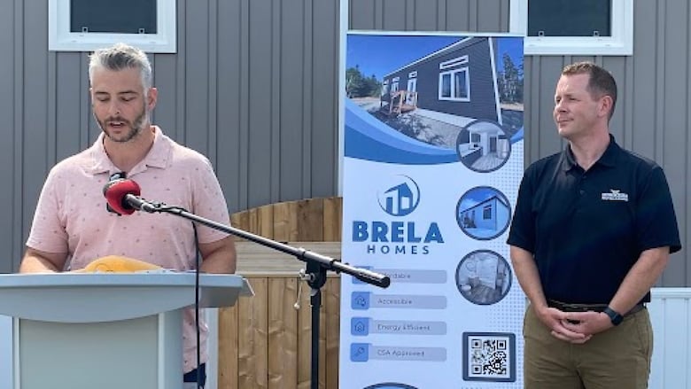A man speaks at a lectern while another man looks on.