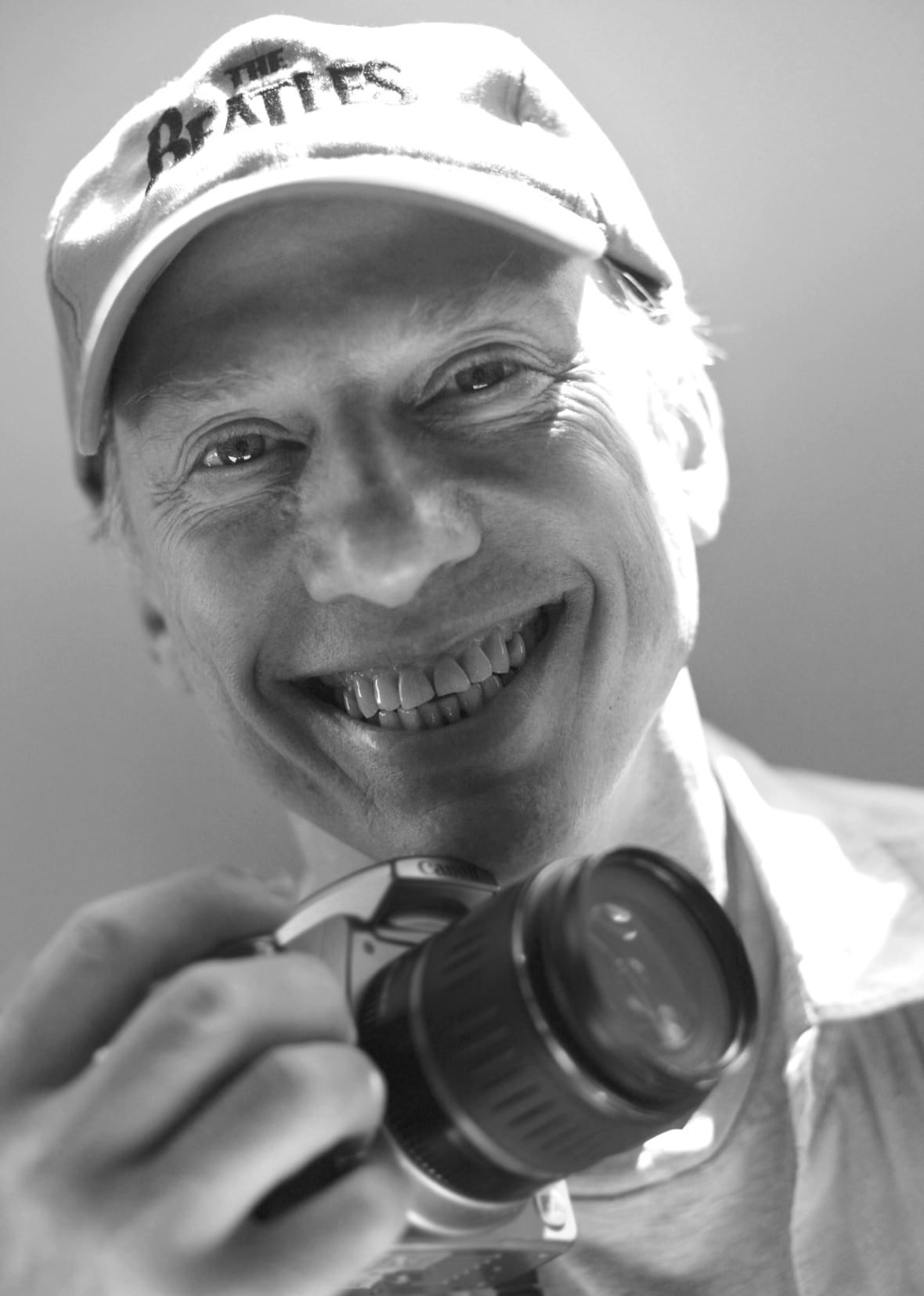 A man in a Beatles baseball cap holds an SLR camera and smiles.