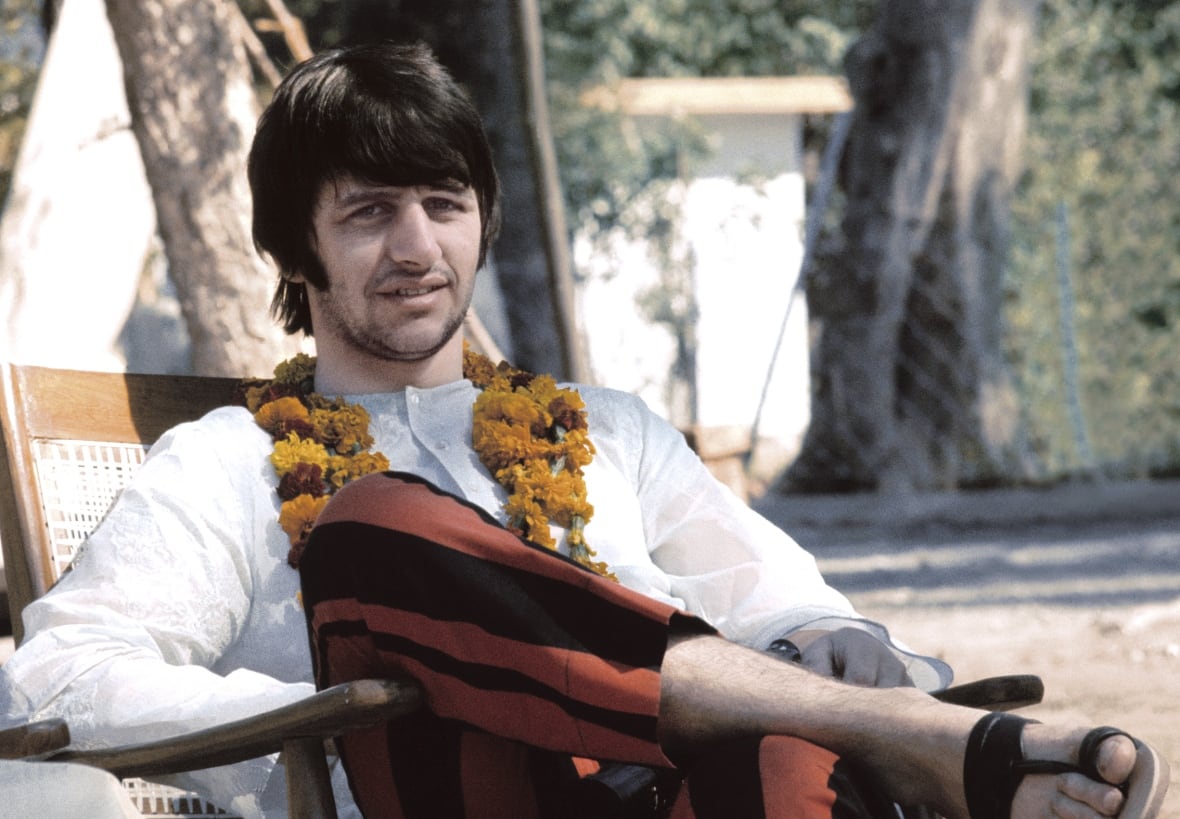 A man in a white shirt and a flower garland sits outside in a chair.