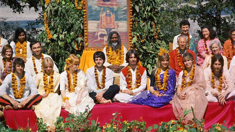 A group of people wearing flower garlands sit cross legged on a platform.