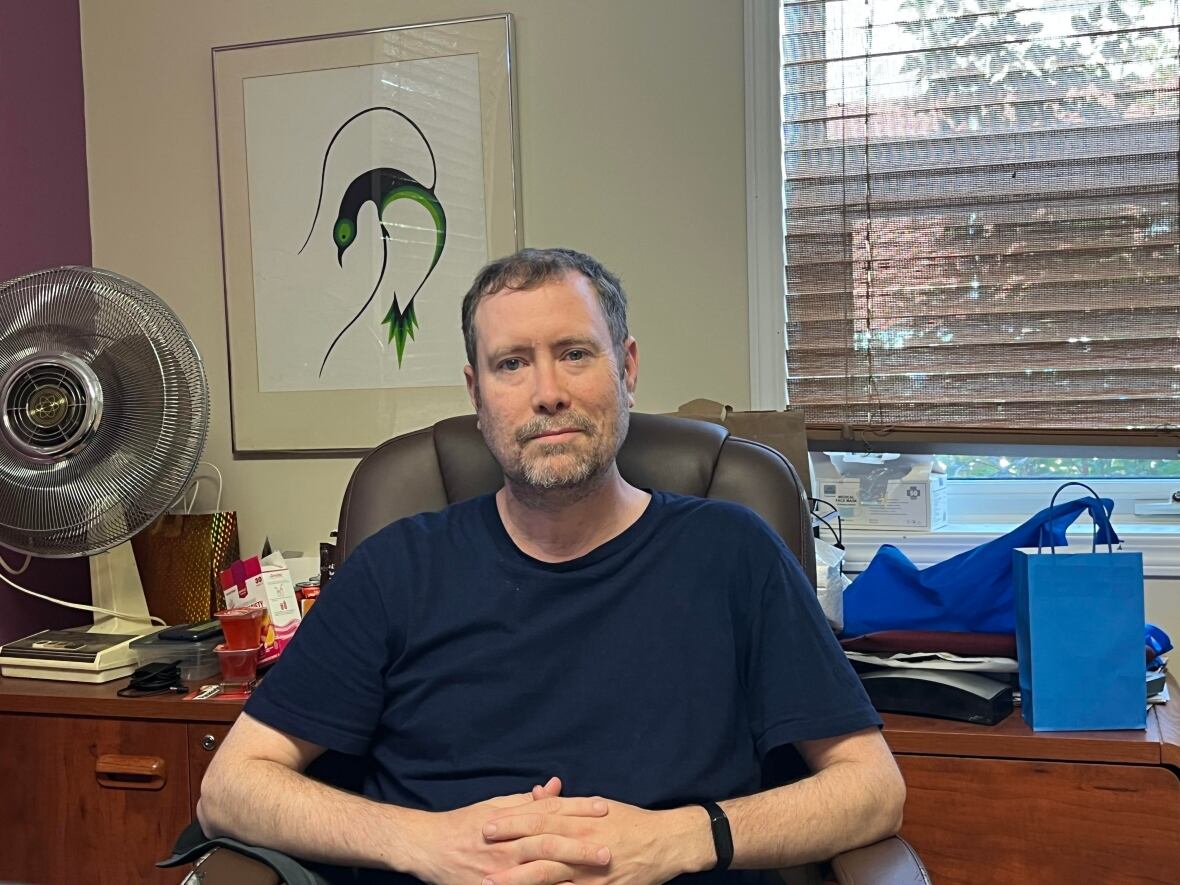 a man in a blue shirt sits with his fingers laced against a beige wall