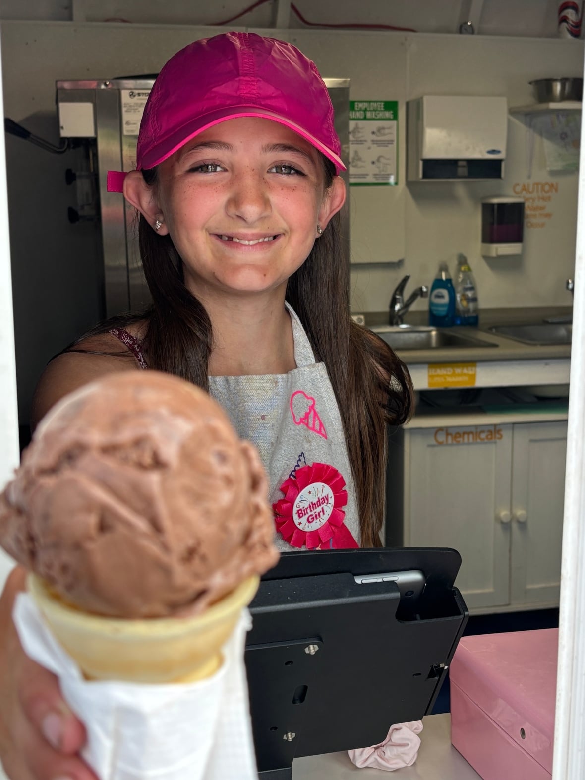 Young girl in pink hat holds ice-cream cone with outstretched hand.