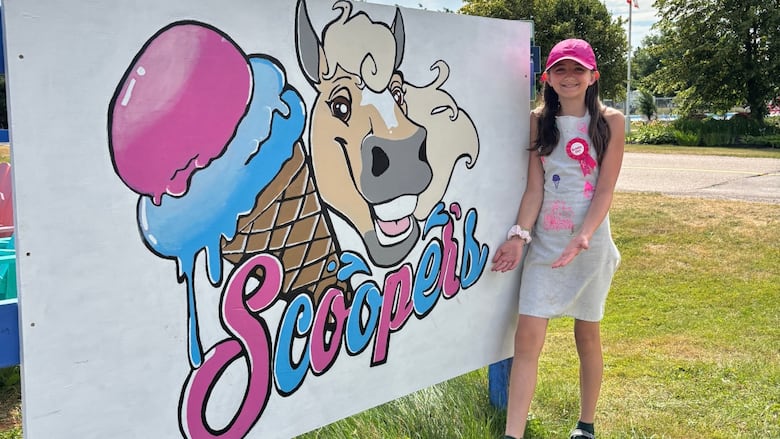 A girl in a pink hat stands with two outreached hands in front of a blue, pink and white sign with the word "scoopers" printed on it.