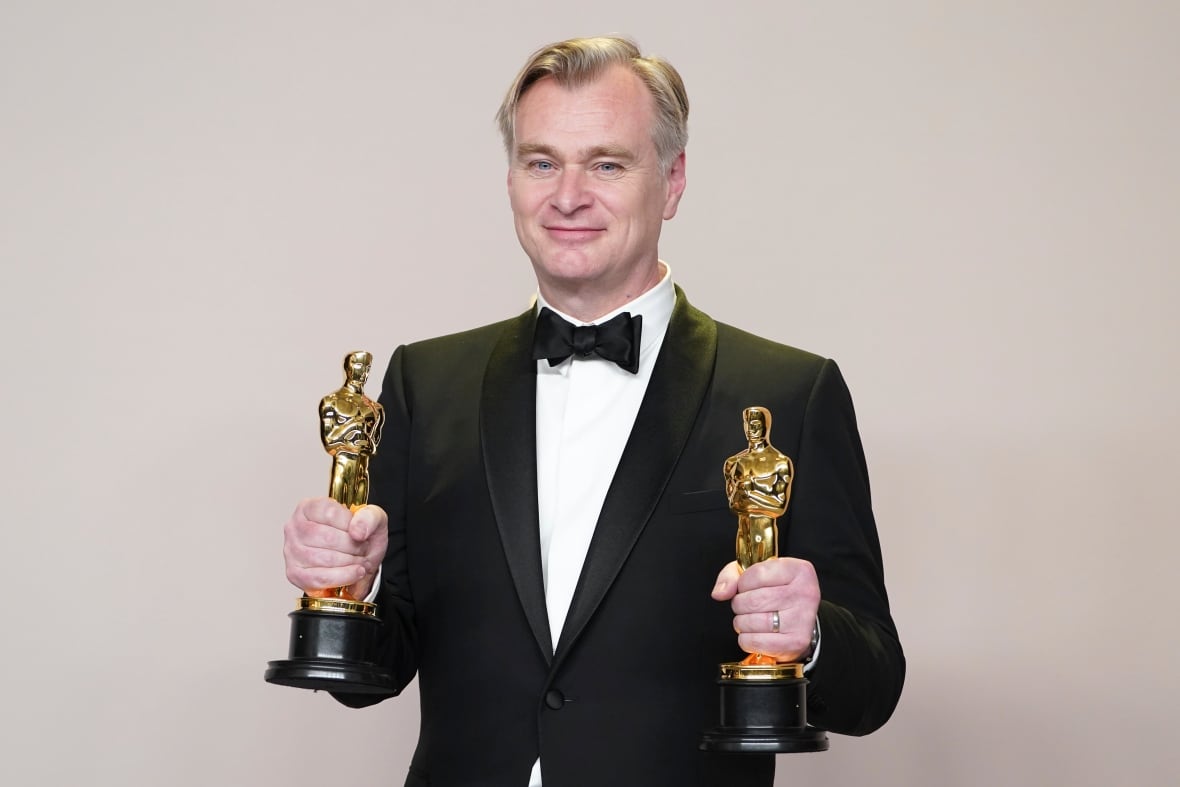 Ein Mann mit grauem Haar, der einen schwarzen Smoking, ein weißes Hemd und eine schwarze Fliege trägt, posiert mit zwei goldenen Oscar-Statuetten in seinen Händen.