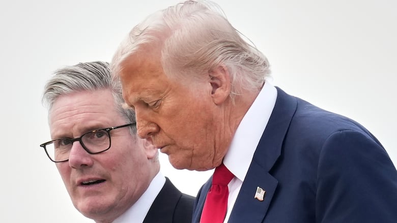 Two men walk side by side against a grey sky. One looks downcast, the other is looking past him to the camera.