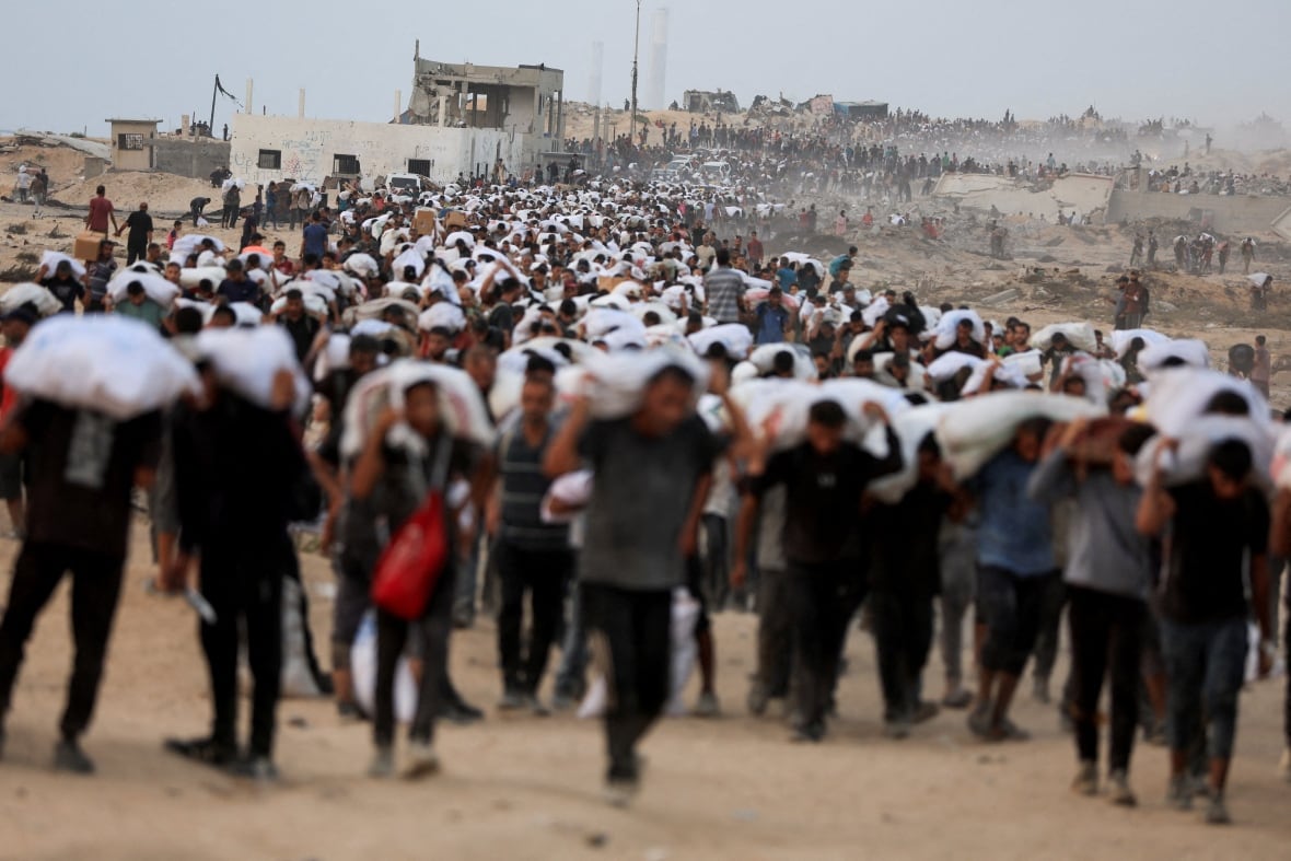 A sea of people walking along a dusty path each carrying a white bag of supplies on their shoulders and backs