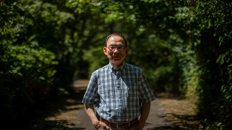 A man wearing a blue plaid shirt and glasses stands among trees on a sunny day