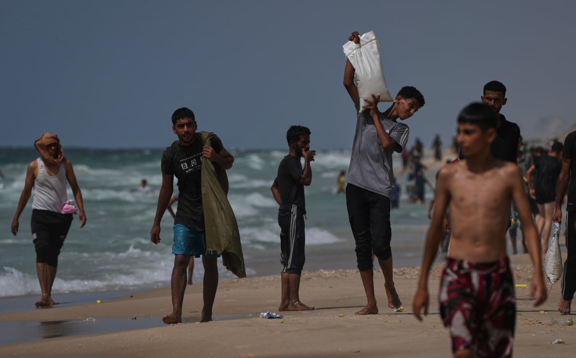 A large group of young men and boys walk along a beach. Some are carrying full, white sacks.