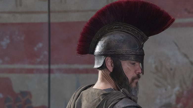 A male actor in costume armor, with a brown cloth cape and silver helmet with a red plume on top, on the set of a movie.