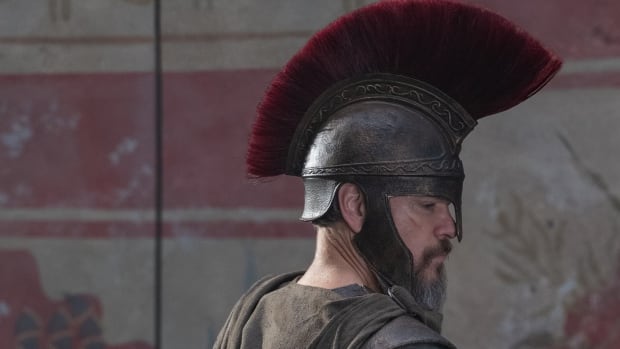 A male actor in costume armor, with a brown cloth cape and silver helmet with a red plume on top, on the set of a movie. 