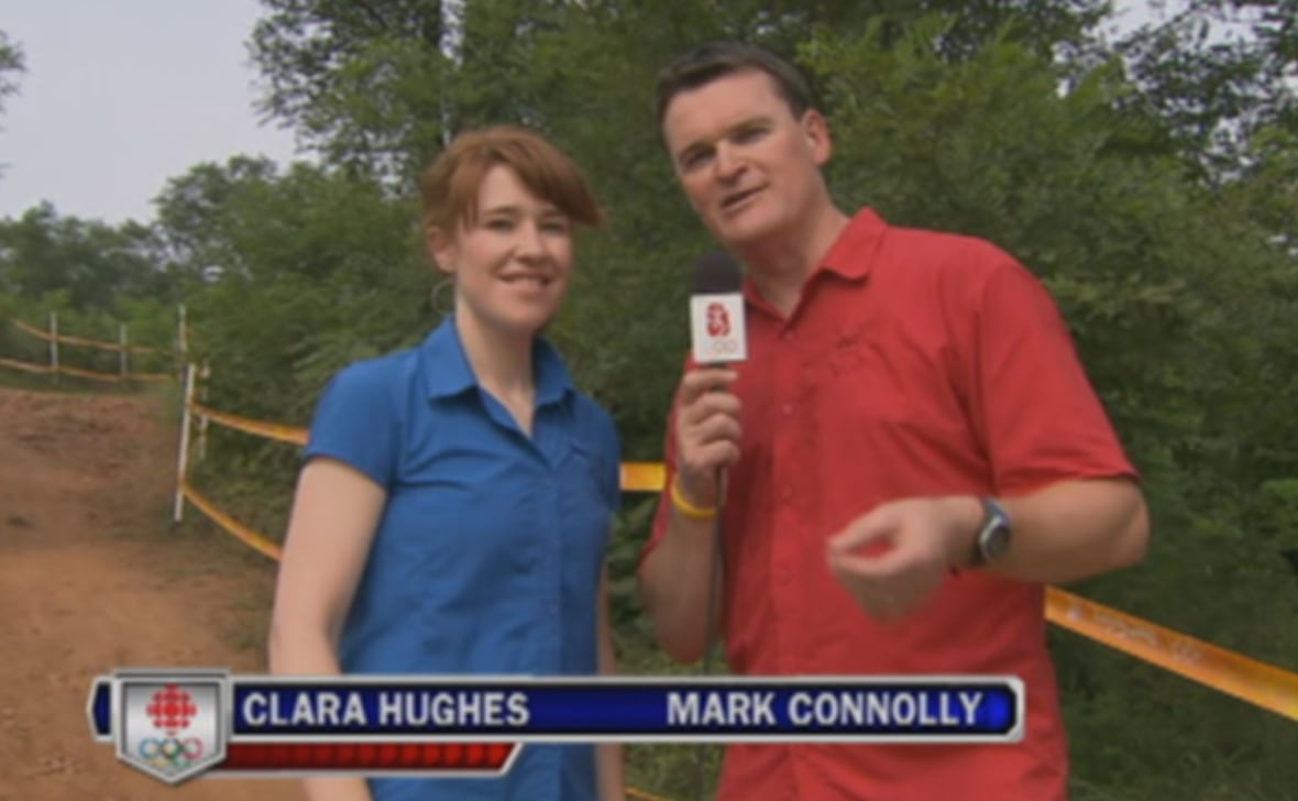 A woman in a blue shirts stands to the left of a man in a red shirt holding a microphone. A dirt bike track going downhill is seen in the background.
