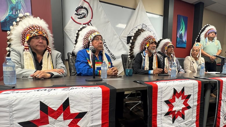 Five men and a woman sit along one side of a table, with microphones placed in front of them. The men are each wearing Indigenous headdresses.