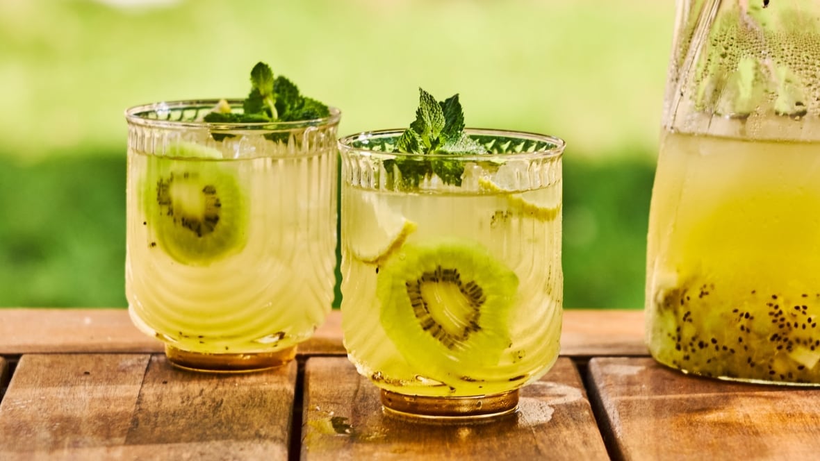 2 glasses of iced green tea with slices of kiwi in them. They're outdoors on a wooden table with the pitcher of the drink sitting next to them.