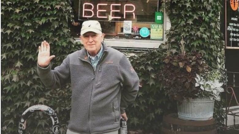 A gray haired man waves in front of a neon "Beer" sign.