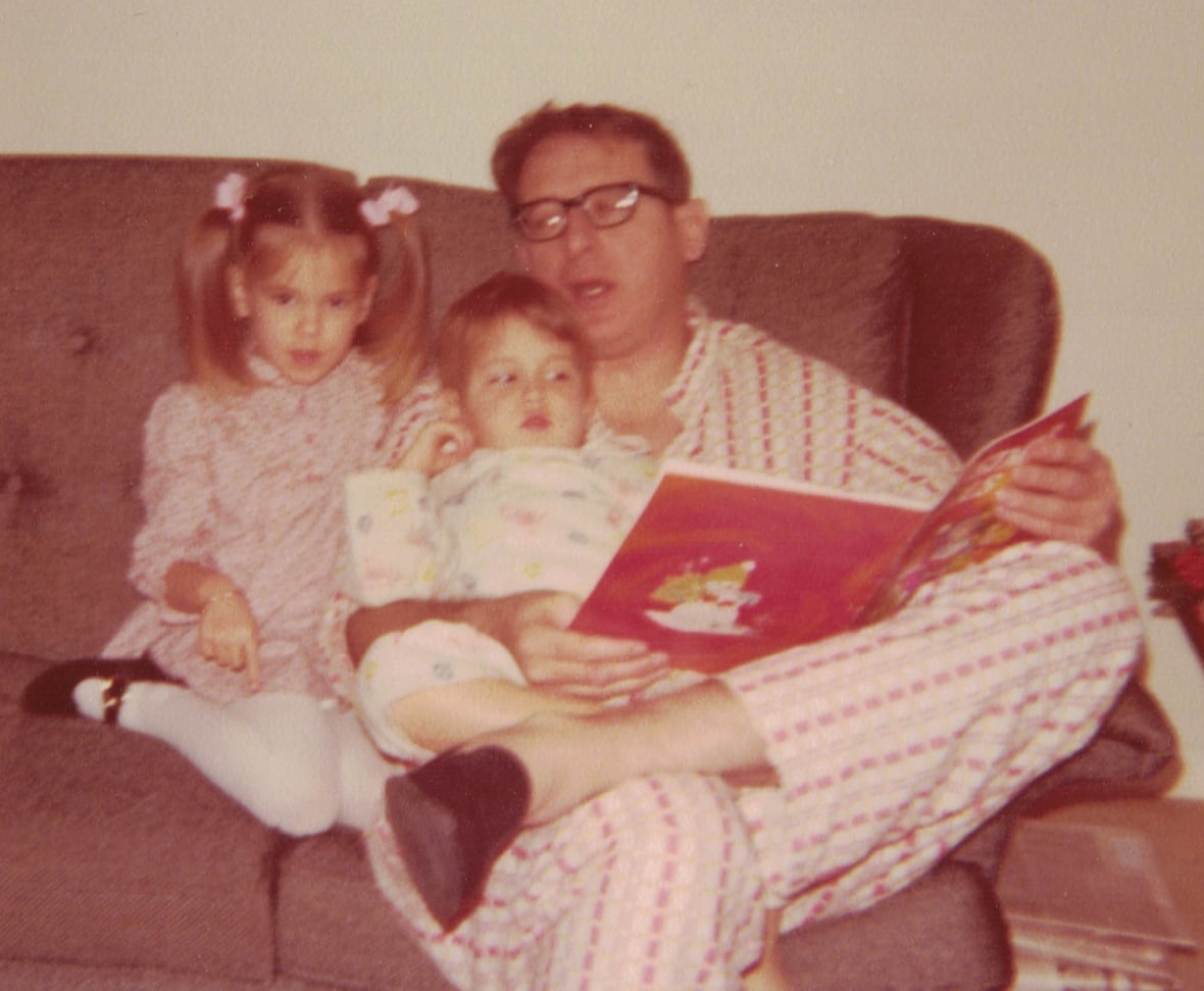 A grainy photograph of a man in pajamas and thick-rimmed glasses sitting on a rose-coloured sofa and reading to a little boy and a little girl. 