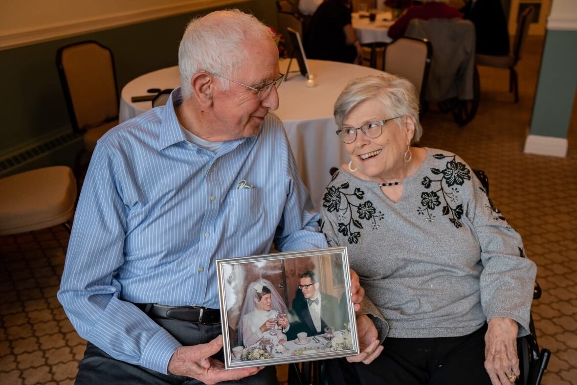 A gray-haired man and woman smile and gaze into each other's eyes. The man is holding their wedding picture. 