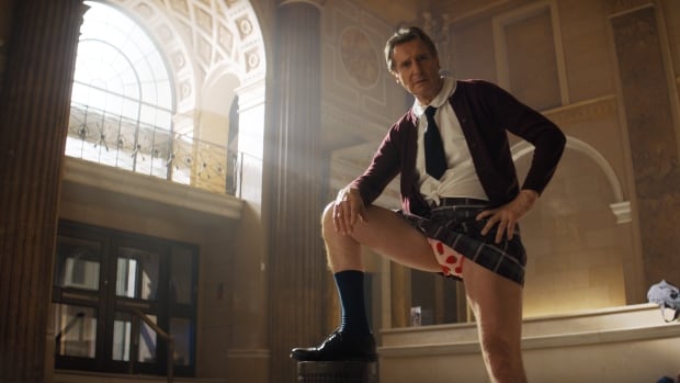 A man wearing an undersized schoolgirl's uniform poses with his foot resting on a pedestal. The skirt he's wearing is riding up, exposing pink underwear. 
