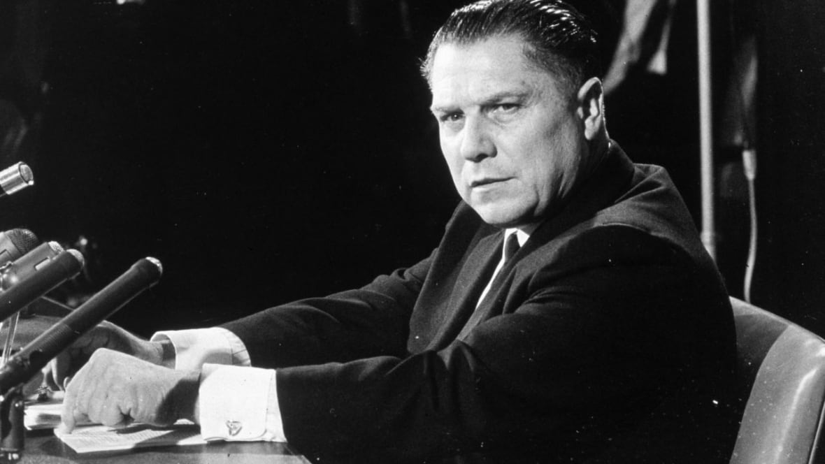 A man in a suit sits in front of microphones.