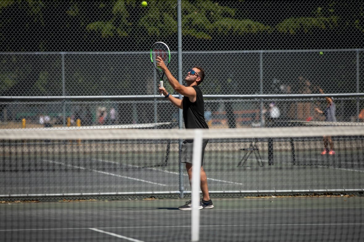 A man in all-black clothing plays tennis in the sun.