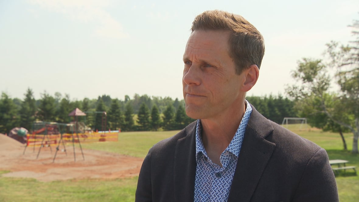 A man in a suit and blue collared shirt stands in front of a playground.
