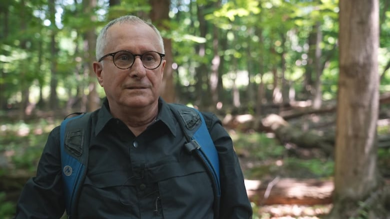 A man wearing a green button up, blue backpack and horn-rimmed glasses looks off the camera.