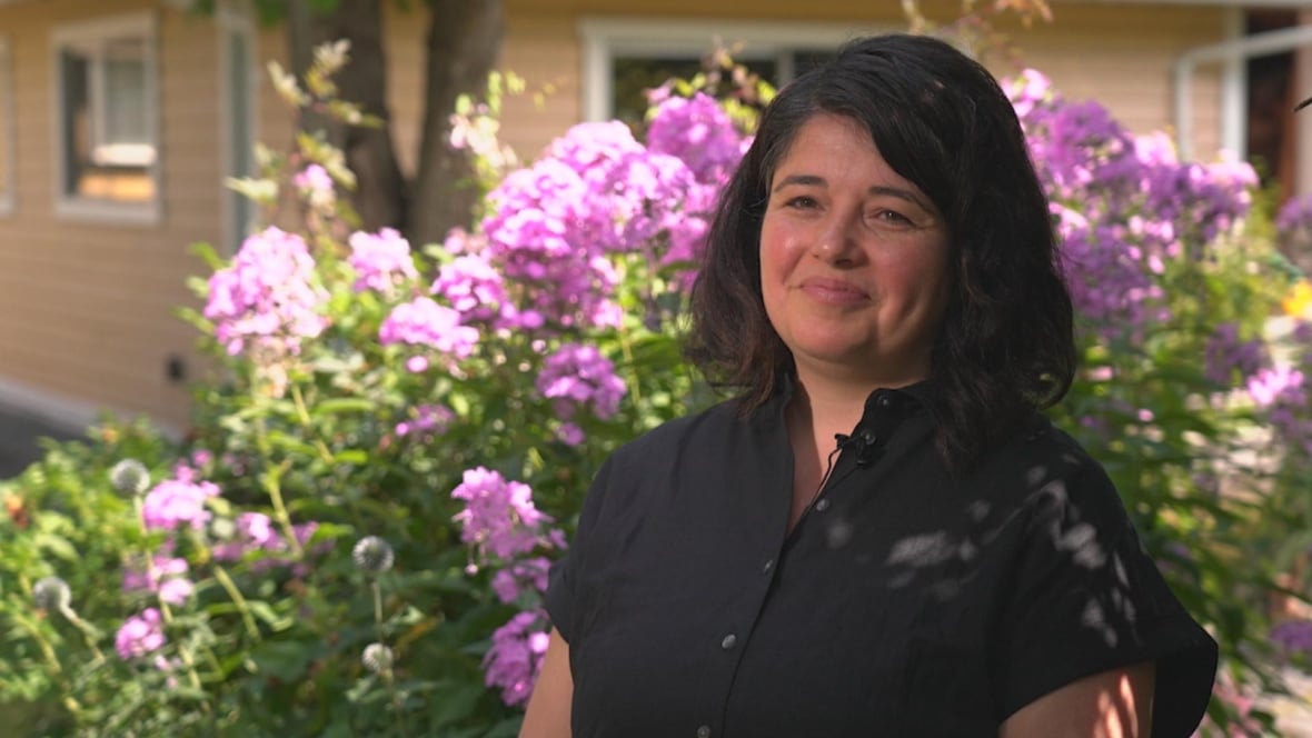 A woman with short hair and a black button down dress smiles slightly at the camera. There is a plant full of purple flowers blooming behind her.