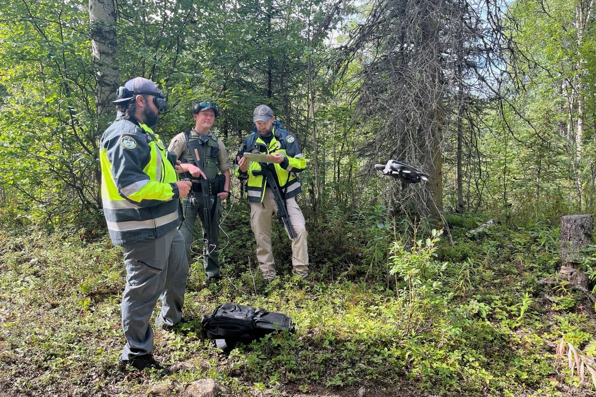 Three men in safety gear and holding firearms stand in a forest.