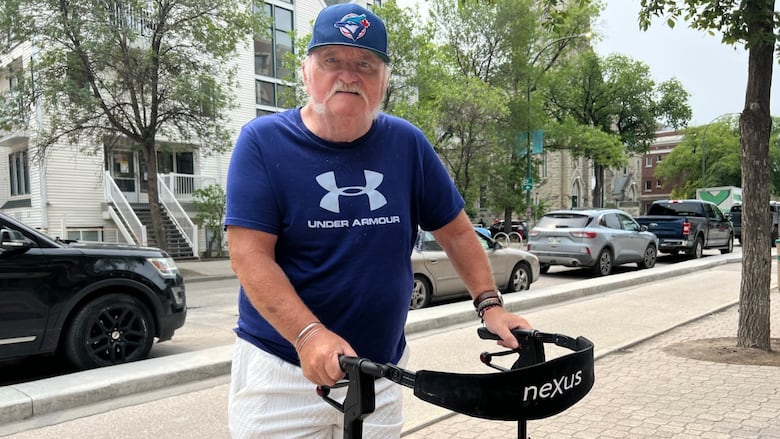 A man with a white mustache, wearing a blue T-shirt, blue baseball cap and white shorts, stands on the sidewalk while holding a walker.