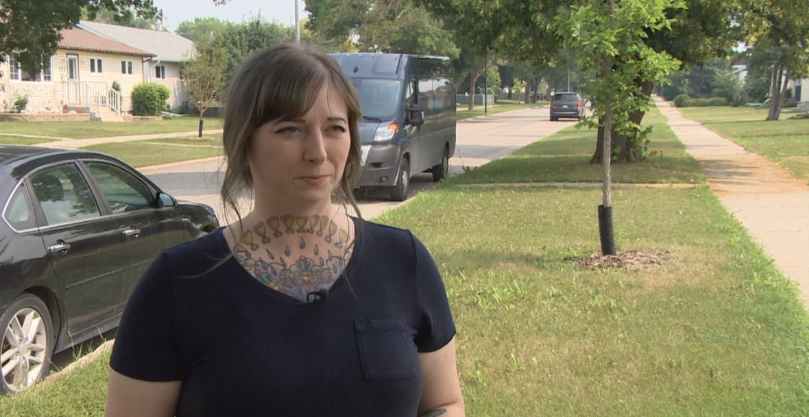 Woman with a chest tattoo and long brown hair pulled back wears a navy blue tshirt. 