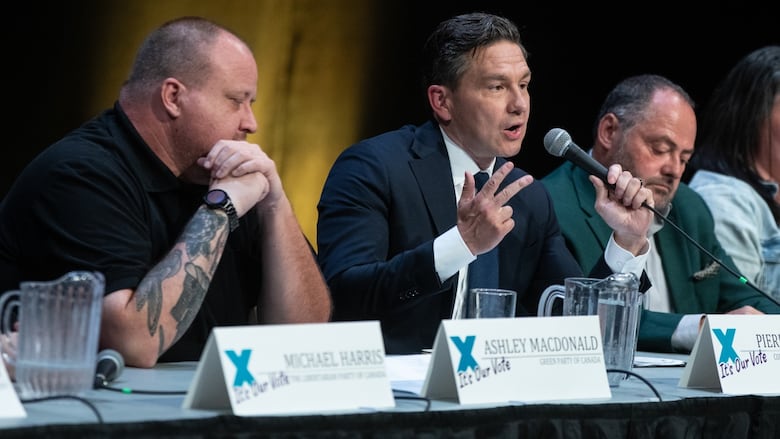 Three men sit at a debate panel, with one holding the mic.