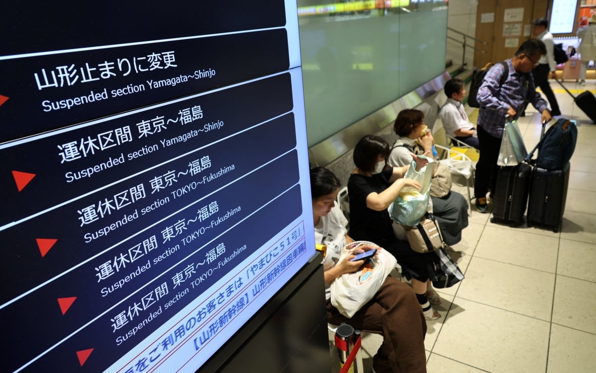 People sit at a train station next to a screen listing out trains temporarily suspended due to the tsunami warning.
