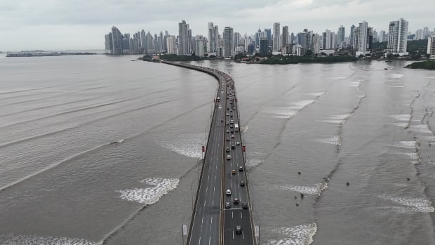 Cars on one side a highway that is over a body of ocean water with wave sets. The road leads to a city skyline.
