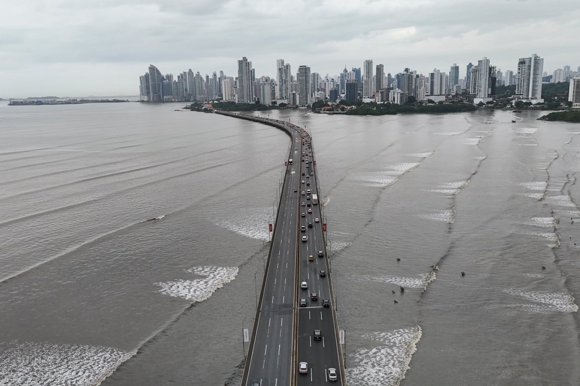 Cars on one side a highway that is over a body of ocean water with wave sets. The road leads to a city skyline.