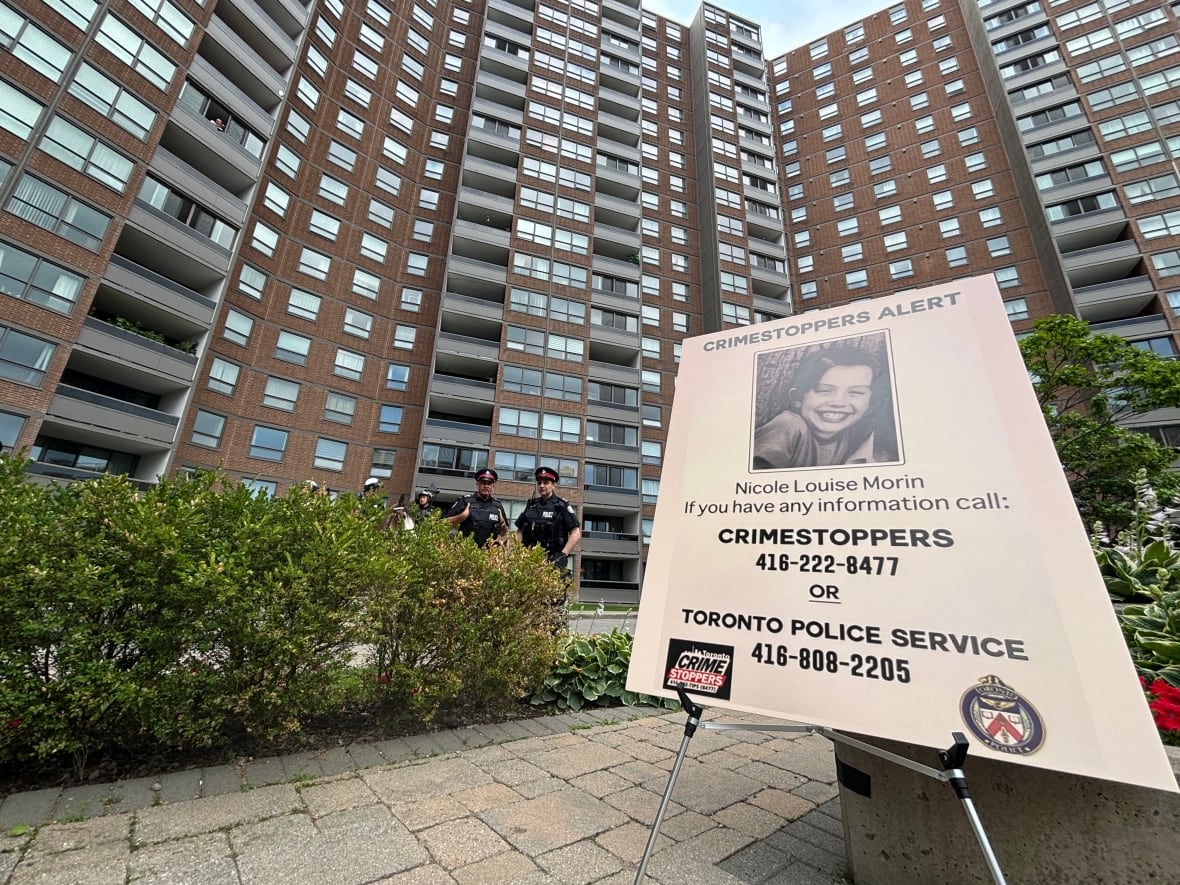 A police poster with a photo of Nicole Louis Morin is pictured with the phone number of police and Crime Stoppers, set up in front of a highrise apartment building with two cops standing in the middle ground.