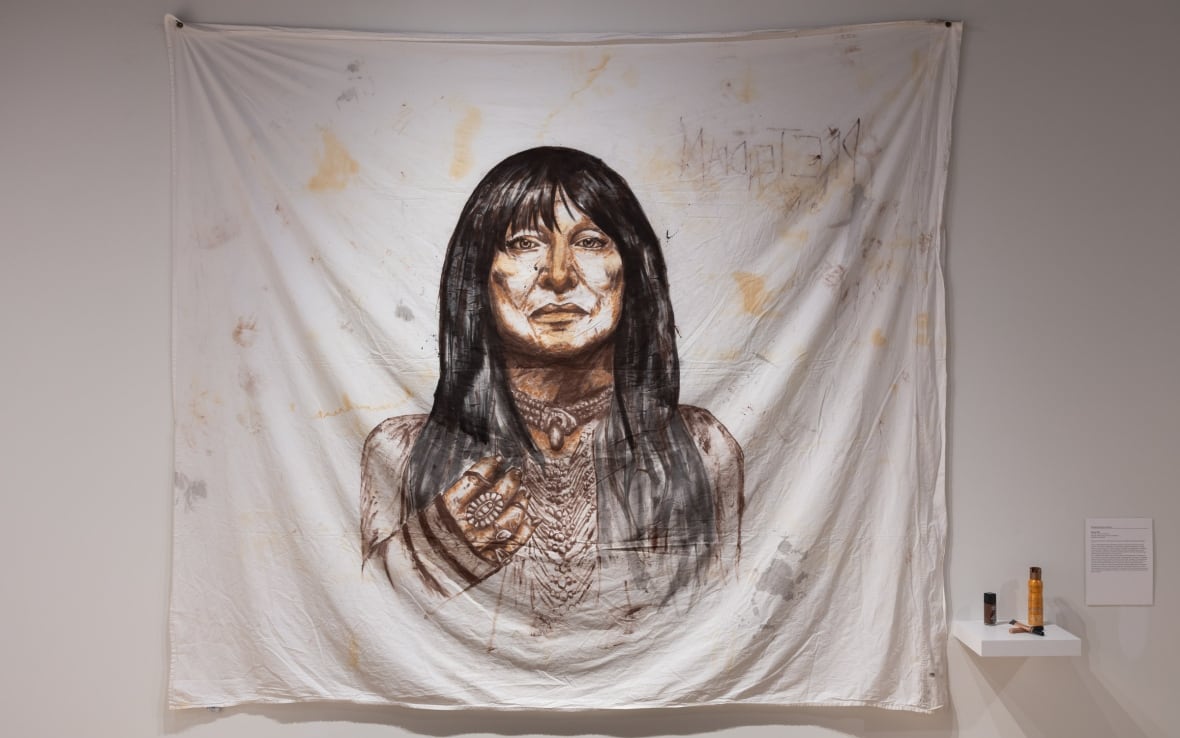 A portrait of Buffy Sainte-Marie drawn in self-tanner, concealer, foundation, and different makeup on a queen-size bed sheet hanging in a gallery.