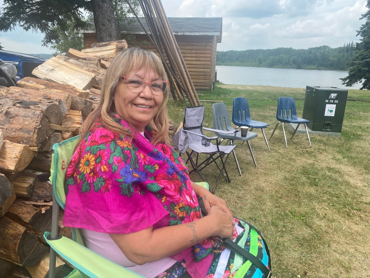 A blonde woman wearing glasses sits at a clearing by a lake, a pink shawl around her shoulders and logs stacked behind her.