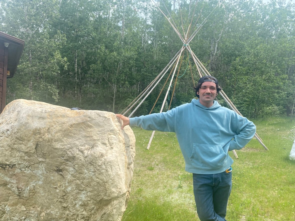 A man in a backwards ballcap and a blue hoodie stands with his arm resting on a giant rock, the structure of a teepee behind him.