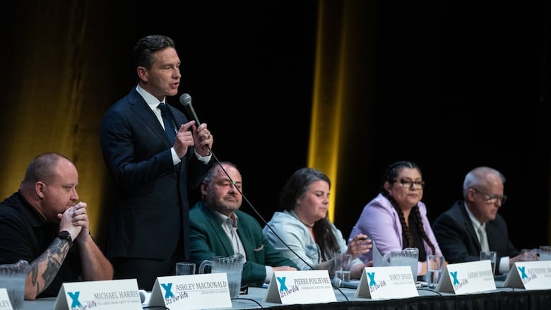 A man stands and speaks into a microphone as a row of people seated beside him listen.