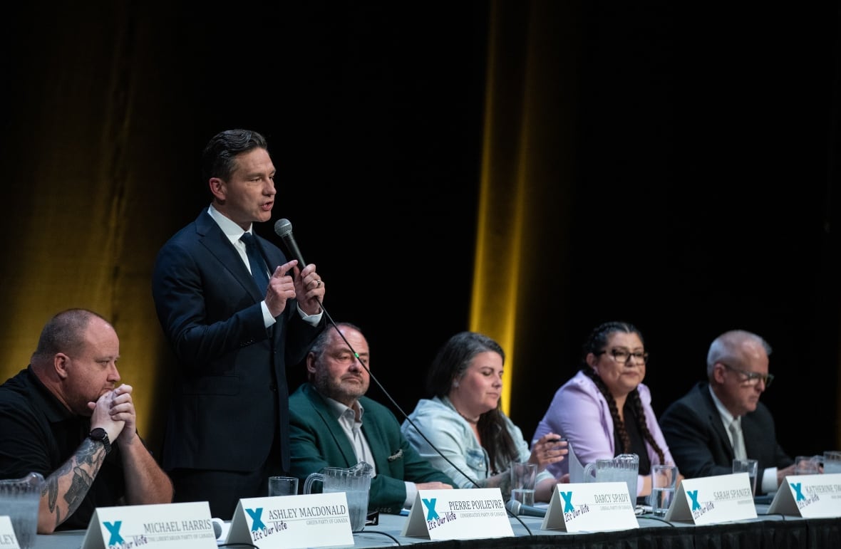 A man stands and speaks into a microphone as a row of people seated beside him listen.