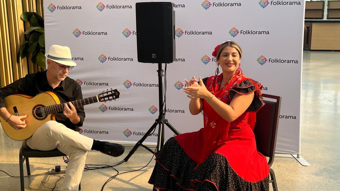 Two people sit on chairs, one playing the guitar, the other clapping. 