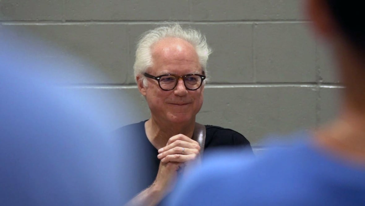 A man with black glasses and thinning hair smiles and holds up folded hands.