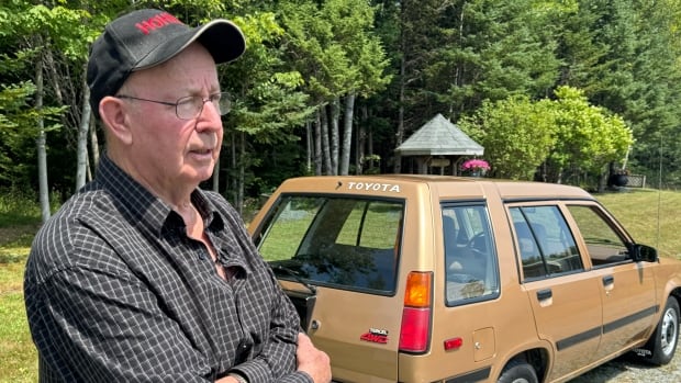 A man in a ball cap and plaid shirt stands with his arms crossed next to a golden-brown car.
