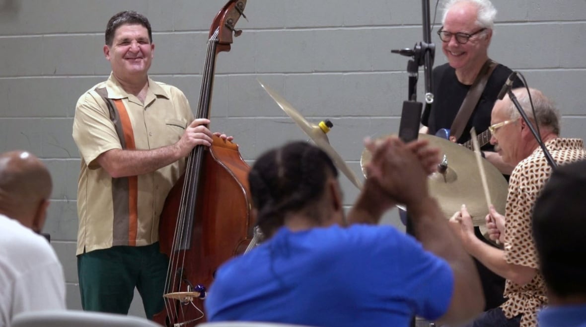 Band members smile while holding their instruments. In the foreground a man in an inmates uniform of blue T-shirt and jeans can be seeing clapping with his hands in the air.