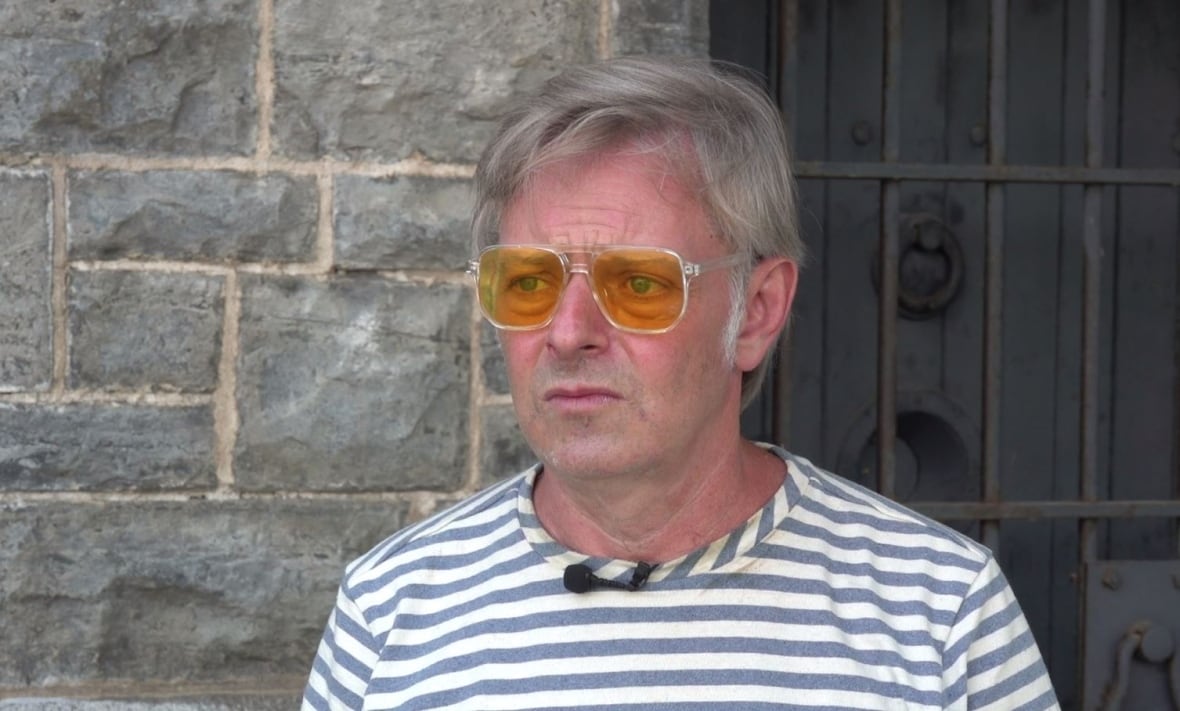 A man with a striped and orange-tined sunglasses stands in front of a stone wall and a barred door.