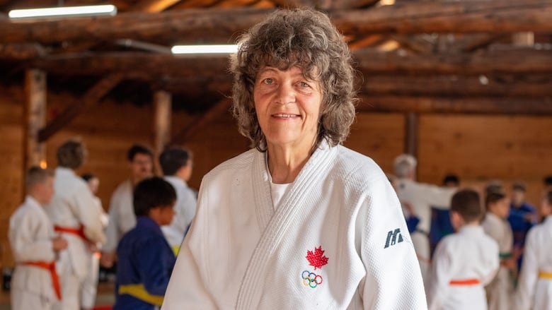 A person in a white judogi is seen standing in a room, slightly smiling.