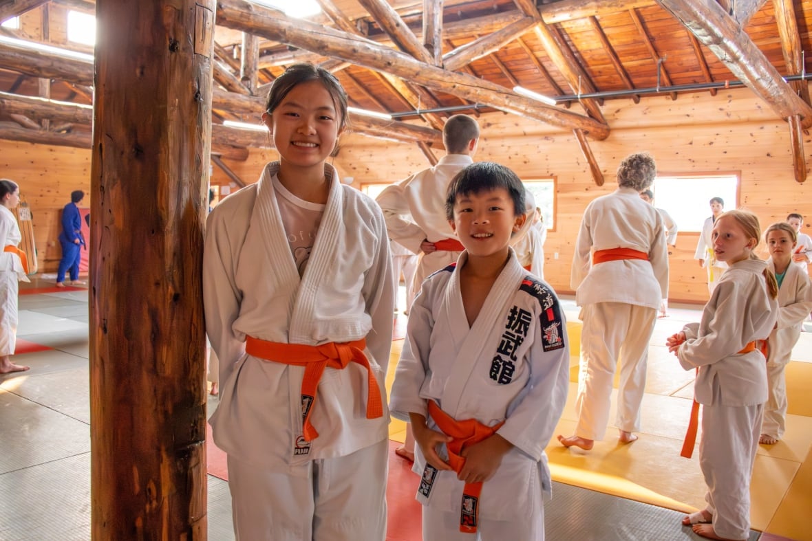 Two people wearing white judogi with orange belts are seen standing indoors by a wooden pole, smiling.
