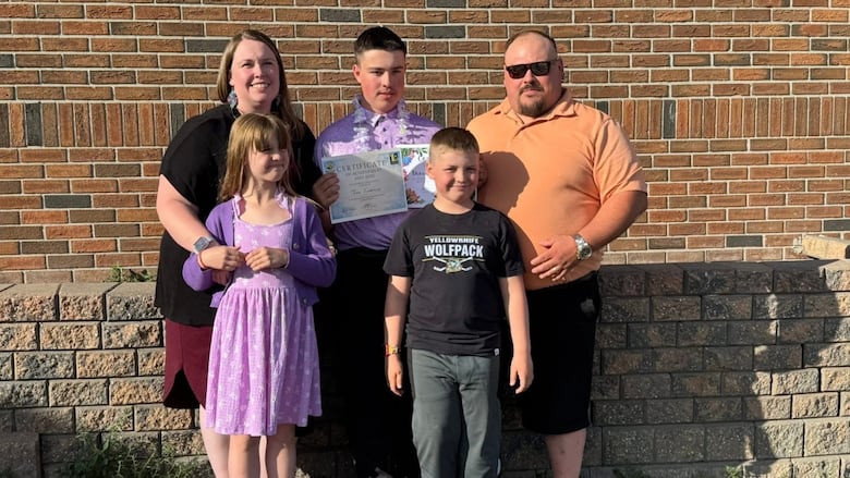 Family stands smiling together, one child holding up a diploma